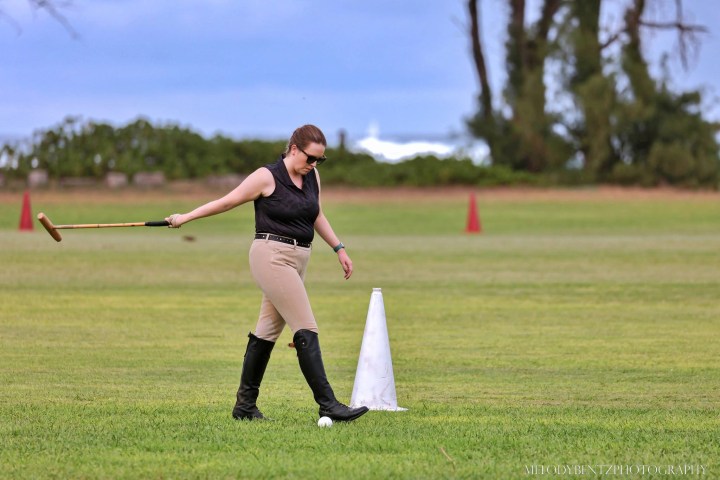 Person walking on a field holding a polo mallet near a cone.