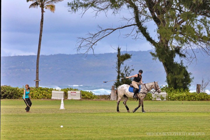 Horse rider playing polo on a field with a person standing nearby, ocean and mountains in background.