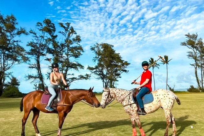 Two people on horseback playing polo on a sunny day with trees in the background.