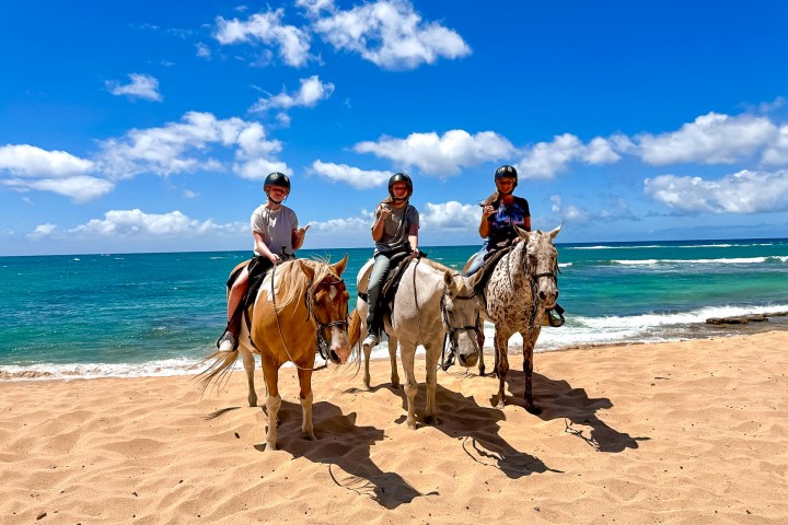 Three people horseback riding on a beach with clear blue skies and ocean waves in the background.