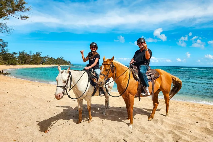 Two people riding horses on a sandy beach beside a blue ocean, waving at the camera under a sunny sky.