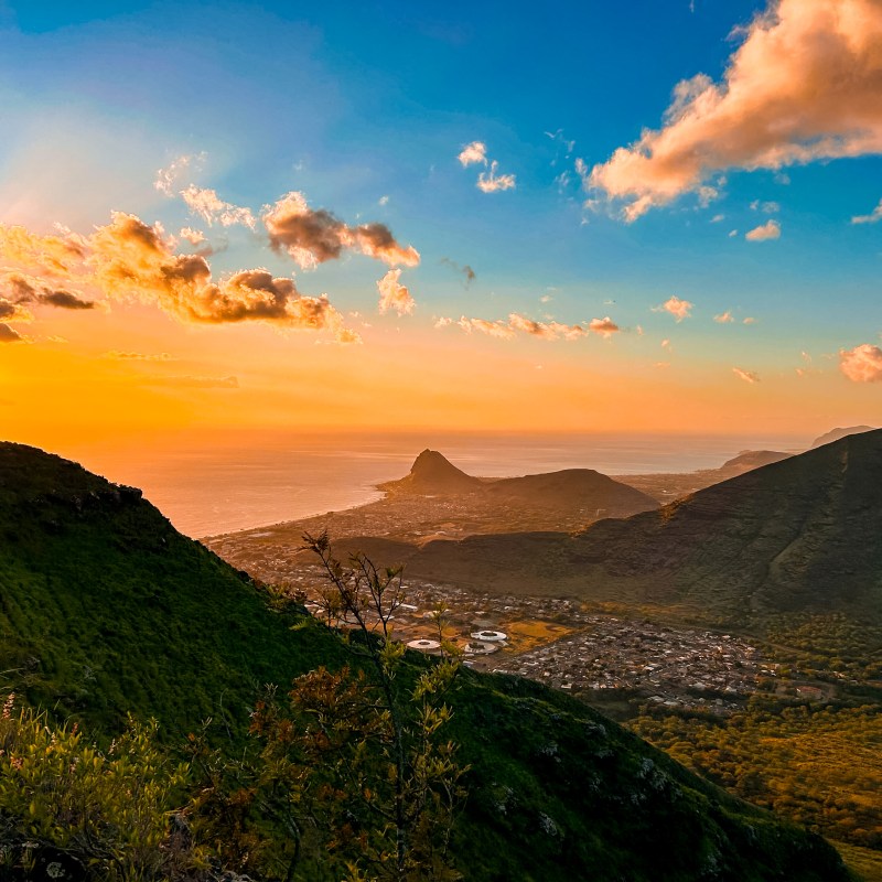 Sunset over a lush green valley with mountains and a town, under a partly cloudy sky.