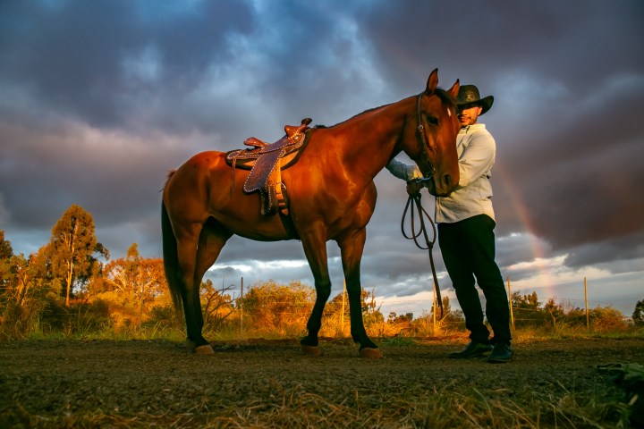 Person in cowboy hat standing with a horse under a cloudy sky with a rainbow.