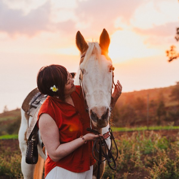 Person in red shirt with a flower in hair lovingly holds a white horse against a sunset background.