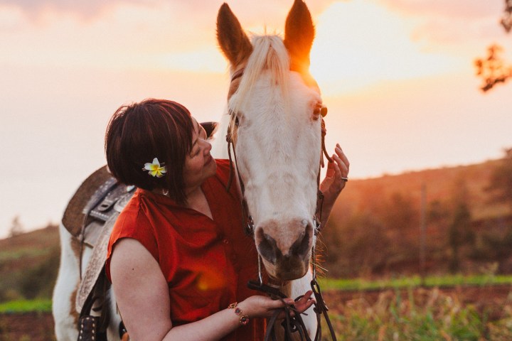 Person in red shirt with a flower in hair lovingly holds a white horse against a sunset background.