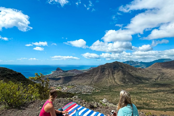 Two people on a picnic blanket with a scenic mountain and ocean view under a blue sky.