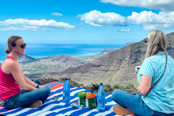 Two people having a picnic on a striped blanket with a mountain and ocean view in the background.