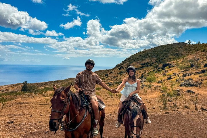 Two people holding hands on horses against a scenic hill and sky backdrop.