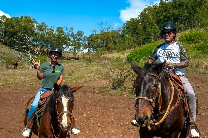 Two people wearing helmets riding horses on a dirt path under a clear blue sky.