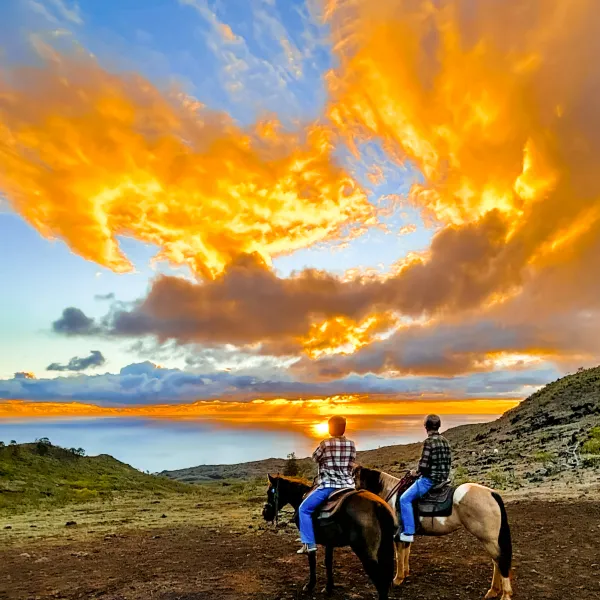 Two people on horses watching a vibrant sunset over a hilly landscape.