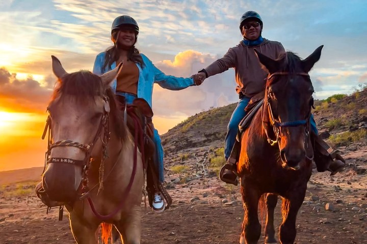Two people on horseback holding hands at sunset on a rocky terrain.
