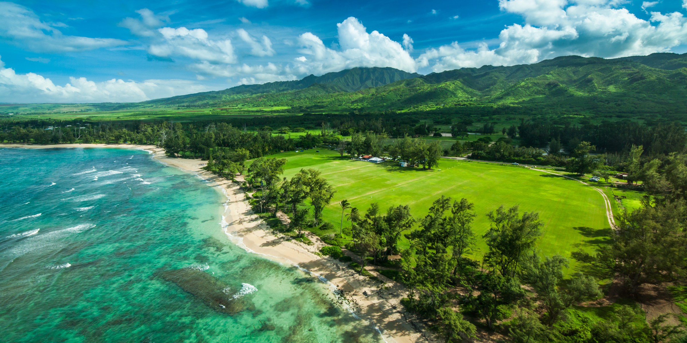 Aerial view of a coastline with turquoise water, sandy beach, green fields, and lush mountains under a blue sky.