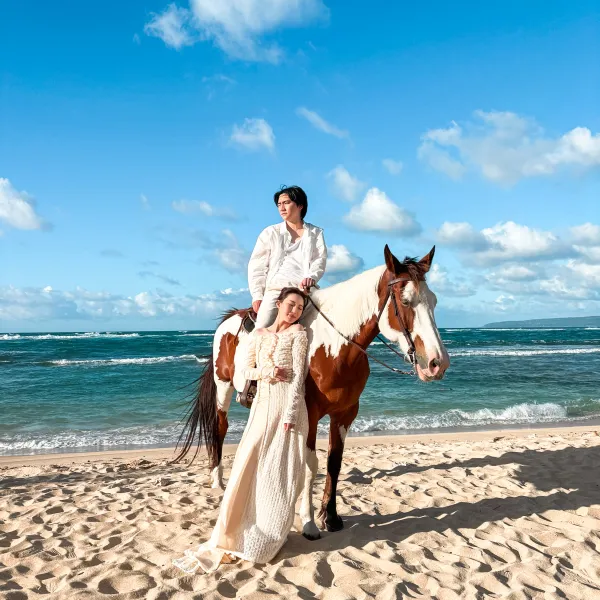 Person in white on horse, another in white standing beside, on a beach under blue sky.