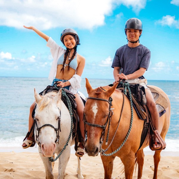 Two people riding horses on a beach, blue sky in background.