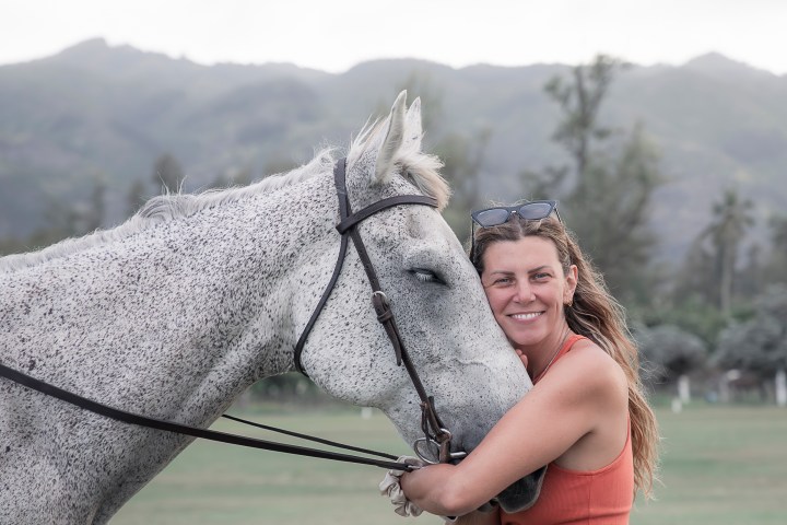Woman in orange tank top hugging a gray horse on a grassy field with mountains in background.