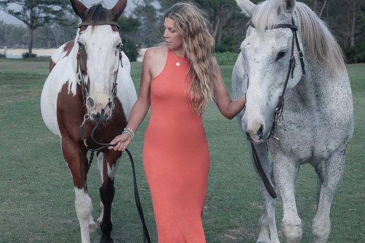 Woman in orange dress walks with two horses on a grassy area, trees in the background.