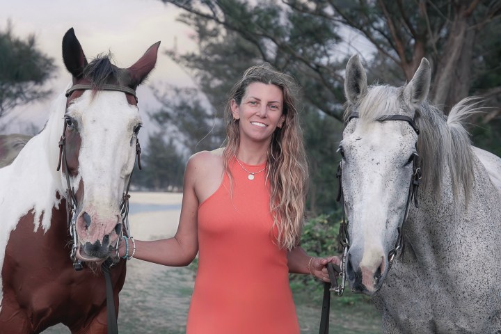 Person in orange dress stands between two horses by trees.