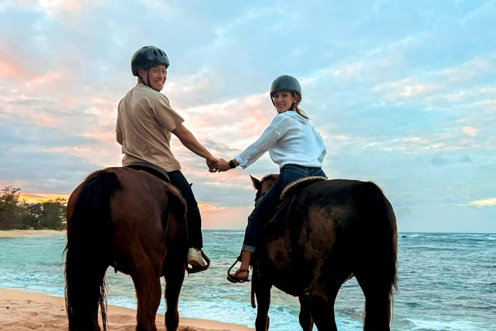 Two people on horseback holding hands on a beach at sunset.
